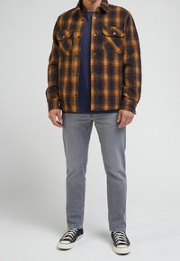 Man in orange and black plaid shirt, navy blue t-shirt, grey jeans, and black sneakers standing against a plain background.