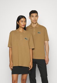 Ruskeat polo-paidat rennolla istuvuudella, joissa on kaksinappinen kaulus ja vasemmassa rinnassa brodeerattu vihreä krokotiililogo.