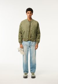 Giacca bomber verde con chiusura a zip, tessuto leggero, abbinata a una camicia verde, jeans dritti blu chiaro e scarpe sportive grigie.