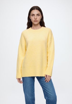 Femme aux longs cheveux foncés portant un pull jaune oversize et un jean bleu, debout devant un fond blanc uni.