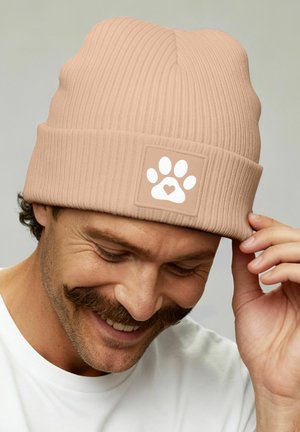 Man smiling while adjusting beige ribbed beanie with white paw print and heart logo, wearing white shirt, neutral background.