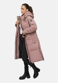 Kvinna i en lång dammig rosa puffjacka med huva, svarta skinny jeans, svarta chunky stövlar, som justerar jackans dragkedja medan hon står på sidan.