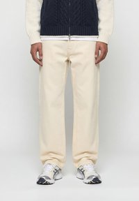 Άτομο που φοράει εκρού παντελόνια με φαρδιά μπατζάκια, λευκά και ασημί αθλητικά παπούτσια, και μπλε πλεκτό γιλέκο με φερμουάρ πάνω από λευκό πουκάμισο με μακριά μανίκια.