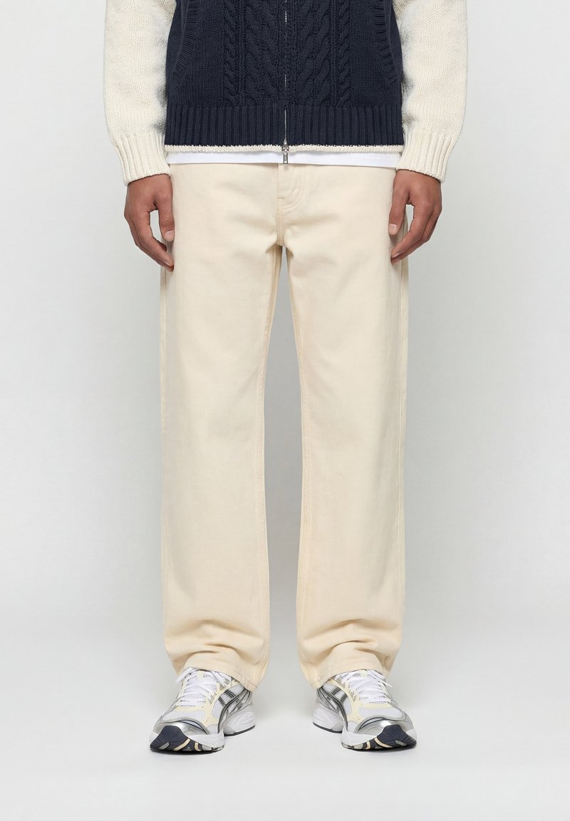 Άτομο που φοράει εκρού παντελόνια με φαρδιά μπατζάκια, λευκά και ασημί αθλητικά παπούτσια, και μπλε πλεκτό γιλέκο με φερμουάρ πάνω από λευκό πουκάμισο με μακριά μανίκια.