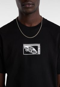 La camiseta negra de algodón presenta un gráfico en blanco de Vans "OFF THE WALL" y un borde de cuadros. Un collar de cadena plateado añade detalle.