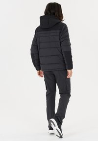 Sort puffjakke med hætte, der har horisontale syninger, kombineret med mørke cargo-bukser og hvide sneakers. Rent design, mat finish.