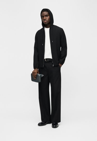 Άνδρας που φοράει μαύρο φούτερ με φερμουάρ, λευκό πουκάμισο, μαύρο παντελόνι φαρδύ και μαύρα παπούτσια, κρατώντας μια μικρή γκρι ορθογώνια τσάντα.