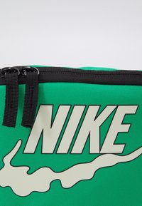 Grön canvasväska med en framträdande vit Nike-logotyp, tecknad i svart, och dubbla dragkedjor upptill. Strukturerad tygyta.