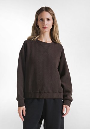 Kvinde med langt blond hår iført en mørkebrun tekstureret sweater og sorte løse bukser, stående mod en ensfarvet hvid baggrund.