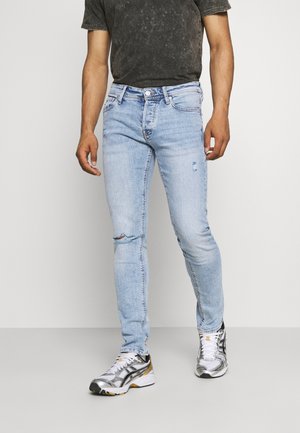 Jeans justos de tom azul claro com detalhes desgastados nos joelhos, combinados com uma t-shirt preta e ténis atléticos prateados.