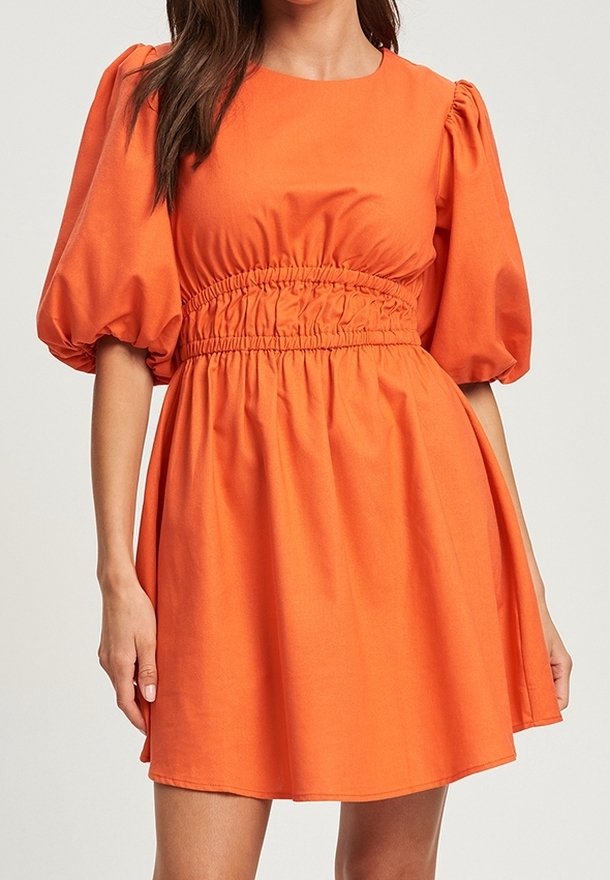 Femme portant une robe orange avec des manches bouffantes jusqu'au coude et une taille élastique froncée, debout devant un fond uni.