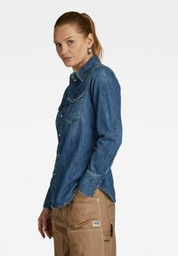 Camicia di jeans blu con maniche lunghe, due tasche sul petto, chiusura con bottoni e cuciture in contrasto. Indossata con pantaloni cargo marroni.
