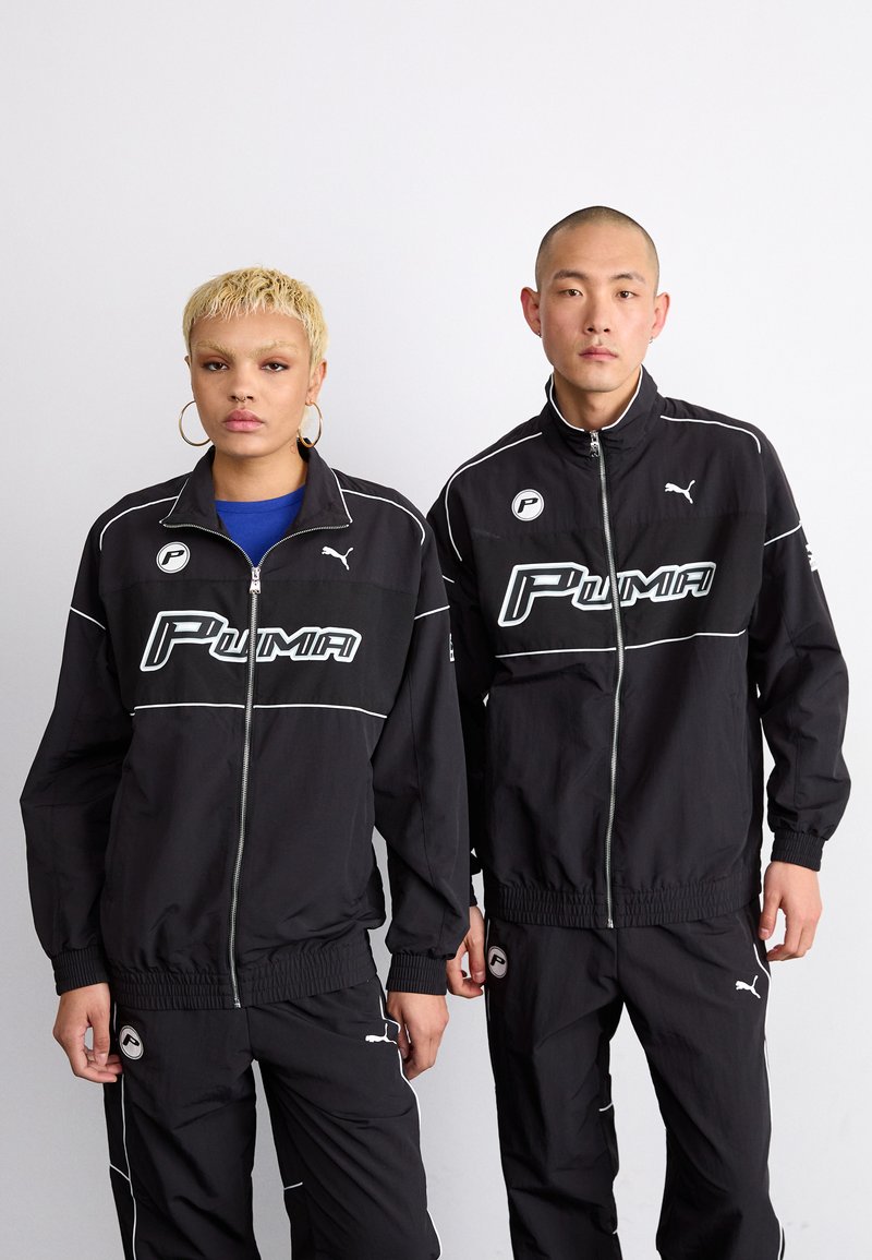 Svart treningsjakker med hvite detaljer og stort "Puma"-logo. Begge har matchende bukser med lignende design og merkevaredetaljer.