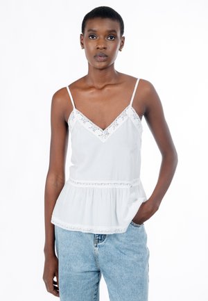 Jeune femme aux cheveux courts portant un haut caraco blanc orné de dentelle et un jean bleu clair, debout avec une main dans la poche.