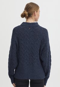 Pull en maille bleu marine avec ourlet côtelé, manches surdimensionnées et motif en forme de diamant texturé. Présente un petit logo à l'arrière du cou.