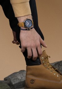 Stivali in pelle marrone con lacci gialli e dettagli neri; orologio da polso con quadrante blu e cinturino beige; mano appoggiata sullo stivale.