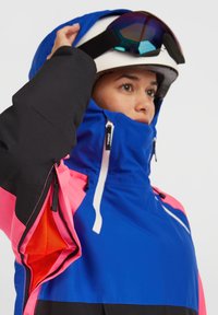 Felblauwe skijas met zwarte en roze accenten, voorzien van een hoge kraag, ritssluitingen voor ventilatie en reflecterende details. Gestructureerde stof.