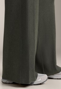 Dunkelgrüne gerippte Hosen mit weitem Bein, die weiße und graue Sneaker bedecken, aufgenommen vom Unterschenkel bis zu den Füßen vor neutralem Hintergrund.