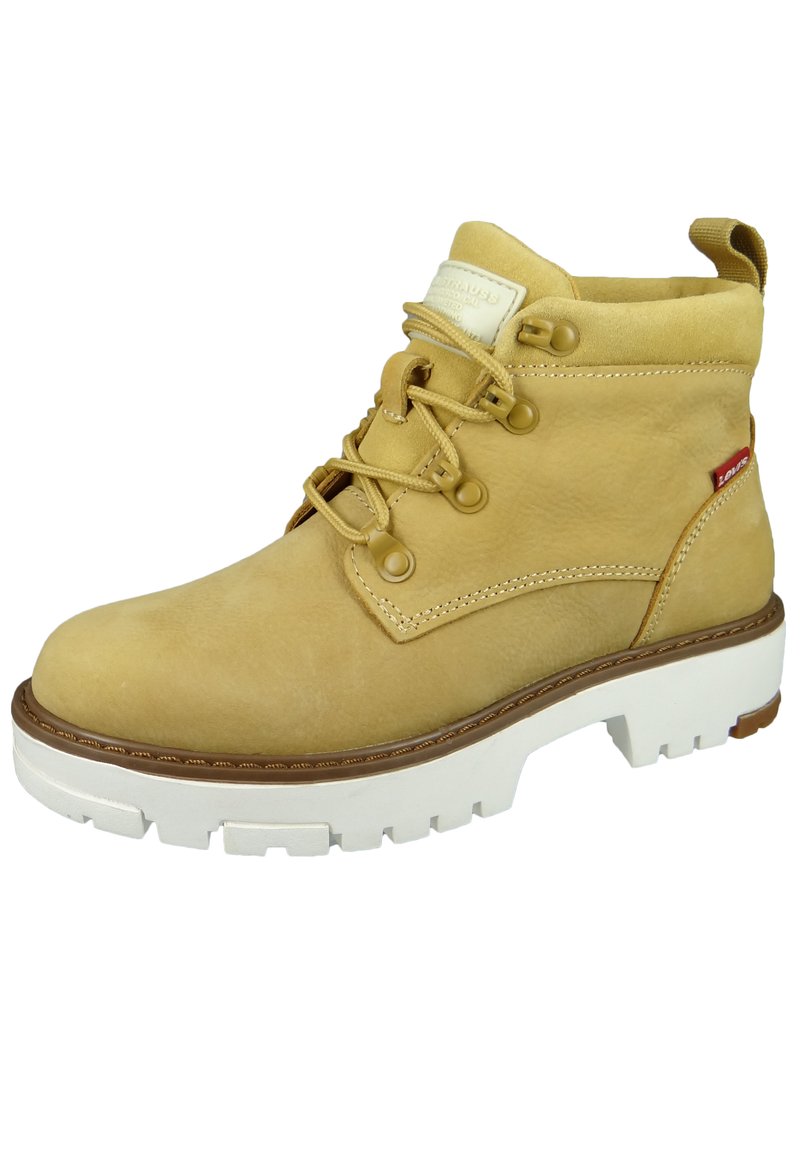 Tan leren enkelboots met een witte chunky zool, voorzien van een vetersluiting, metalen oogjes, een trekband en een rode logo-accent aan de zijkant.