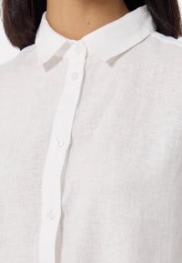 Camicia bianca in lino con colletto classico, dotata di bottoni in plastica trasparente e un motivo di tessuto testurizzato. Dettaglio in primo piano.