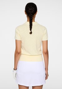 Keltainen golfpaita kauluksella, jossa on teksturoitu kuvio, lyhyet hihat ja istuva vyötärö, yhdistettynä valkoiseen golfhameeseen.