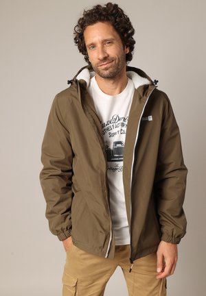 Man met krullend haar draagt een bruine jas over een witte T-shirt met print en beige cargobroek, staat met handen in de zakken tegen een effen achtergrond.