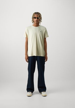 T-shirt en coton jaune clair avec un petit logo, associé à un jean large bleu foncé et des baskets blanches. Design simple et décontracté.