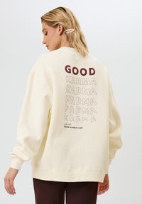 Felpa color crema con grafiche "GOOD" e "KARMA" in rosso e beige sul retro; vestibilità comoda con polsini e orlo a costine.