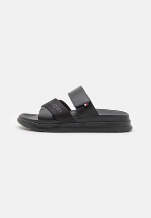 Sandale noire à bout ouvert avec deux brides, une bride en tissu avec le texte "HILFIGER" et une bride en cuir velcro réglable avec un petit drapeau rouge, blanc, bleu.