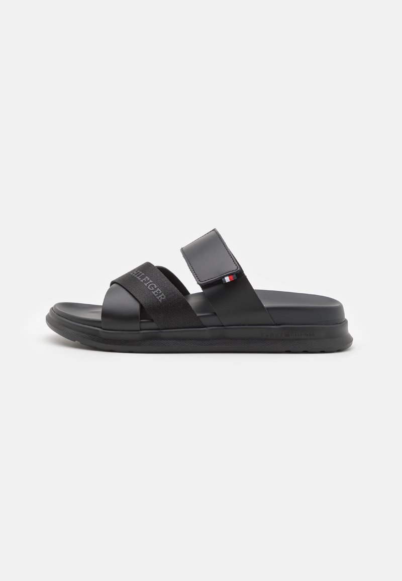 Sandale noire à bout ouvert avec deux brides, une bride en tissu avec le texte "HILFIGER" et une bride en cuir velcro réglable avec un petit drapeau rouge, blanc, bleu.