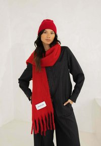 Rode wollen sjaal met franjes en een label, gedragen over een zwart overhemd met een subtiele krijtstreep. Het model draagt ook een bijpassende rode muts.