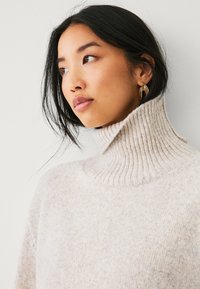 Maglione a collo alto color crema con una texture a coste e una vestibilità morbida. Presenta un colletto alto e un materiale lavorato a maglia morbido. Design semplice e neutro.