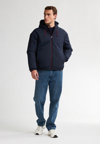 Jachetă puffer bleumarin cu glugă, cu accente roșii și fermoar frontal. Asociată cu blugi albaștri și adidași albi pe un fundal simplu.