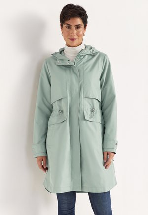 Kvinde iført lys grøn hættejakke over hvid rullekrave og blå jeans, stående mod en ensfarvet hvid baggrund.