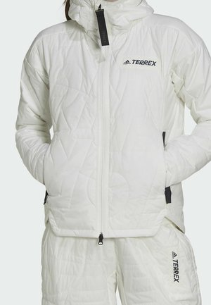 Chaqueta y pantalones blancos acolchados Adidas Terrex con cremallera frontal y bolsillos laterales, manos en los bolsillos de la chaqueta, logotipos de la marca visibles en el pecho y la pierna.