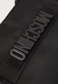 Mustast kangast kott, millel on tekstuuriga pind, lai rihm, millel on kõrglõikega logo, ja kontrastsed mustad aksessuaarid. Vastupidav disain ja puhtad jooned.