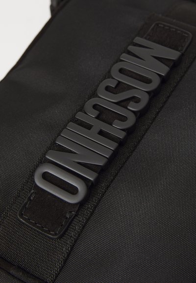 Svart tygväska med texturerad yta, bred rem med präglad logotyp och kontrasterande svarta detaljer. Robust design och rena linjer.