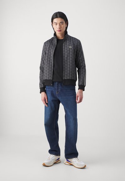 Veste bomber noire à motifs avec un zip sur le devant, poignets côtelés, portée sur une chemise noire, associée à un jean bleu et des baskets blanches.
