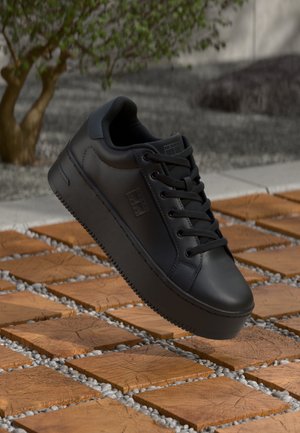 Zapatillas negras de cuero liso con punta redonda, diseño con cordones, suela plana y detalles texturizados. El fondo presenta baldosas marrones y piedras.