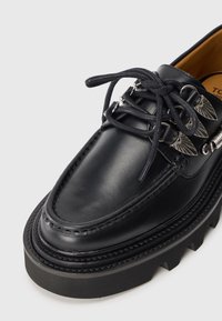 Chaussure noire en cuir à lacets avec une semelle en caoutchouc épaisse, présentant des accents en métal argenté et un bout cousu dans le style mocassin.