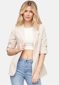 Beige, getailleerde blazer met een notch-kraag en opgerolde mouwen. Soepele stof; gecombineerd met een witte crop top en lichtblauwe high-waisted jeans.