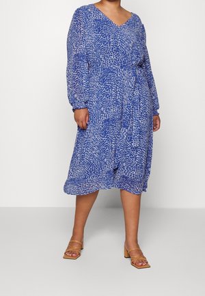 Vrouw draagt een midi-jurk met blauwe en witte print, lange mouwen, en een getailleerde taille, gecombineerd met beige sandalen met hak.