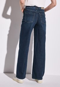 Pantalones vaqueros de mezclilla oscura de pierna ancha con cintura alta, dos bolsillos traseros y un acabado limpio en los dobladillos. Combinados con zapatillas blancas.