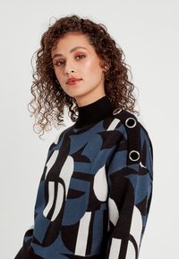 Maglione a motivi blu, nero e bianco con colletto alto, dotato di dettagli metallici circolari sulle spalle e tessuto a maglia testurizzato.