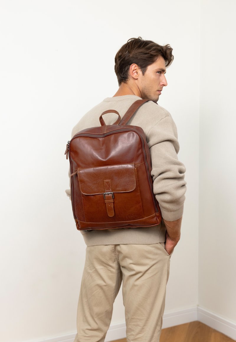 Mann in beiger Sweater und Hose mit braunem Leder-Rucksack, der an einer weißen Wand steht und mit einer Hand in der Tasche steckt.