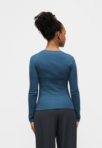 Γυναίκα με σγουρά μαλλιά μαζεμένα σε κότσο, που φοράει ένα εφαρμοστό μπλε πλεκτό τοπ με μακριά μανίκια και σκούρο γκρι παντελόνι, εμφανιζόμενη από πίσω σε απλό φόντο.