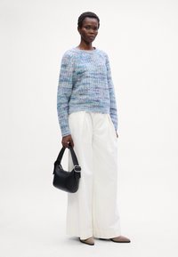 Gestrickter Pullover in sanften Blau-, Lila- und Grüntönen; lockere Passform. Weiße weit geschnittene Hosen. Schwarze Handtasche mit strukturierter Form, über der Schulter getragen.