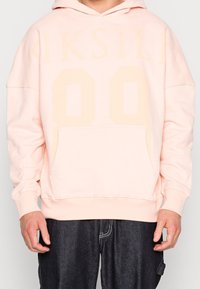 Jasnoróżowa bluza z kapturem z dużym, centralnym nadrukiem "00", przednią kieszenią i sznurkiem do ściągania kaptura. Miękka tkanina o luźnym kroju.