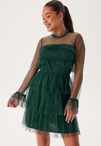 Robe verte à volants en tissu transparent avec des pois, dotée de longues manches transparentes, de volants et d'une taille cintrée.