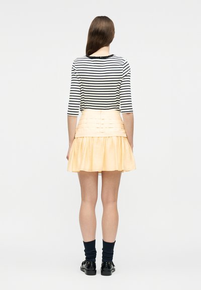 Γυναίκα που στέκεται με την πλάτη προς το θεατή, φοράει ριγέ μπλούζα σε μαύρο και άσπρο, ανοιχτό ροδακινί πλισέ φούστα, μαύρες κάλτσες και μαύρα παπούτσια, με απλό φόντο.